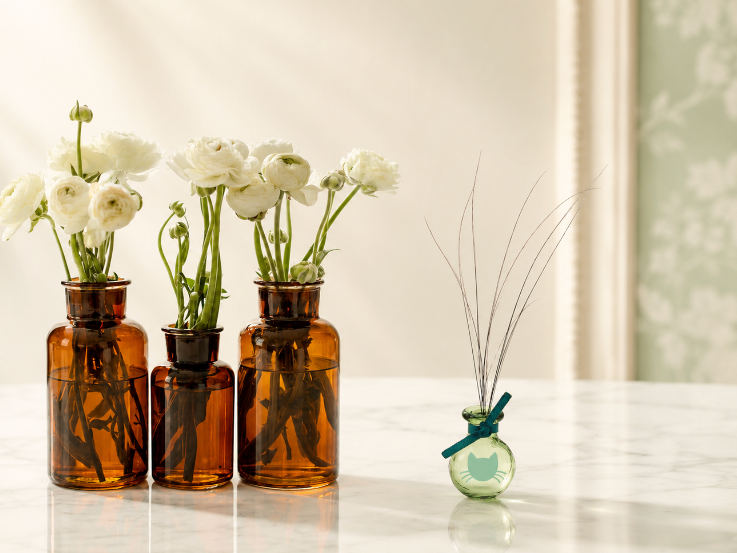 Tiny green glass Whisker Vase holding fine cat whiskers beside amber bottles of white flowers on a marble surface with soft light.