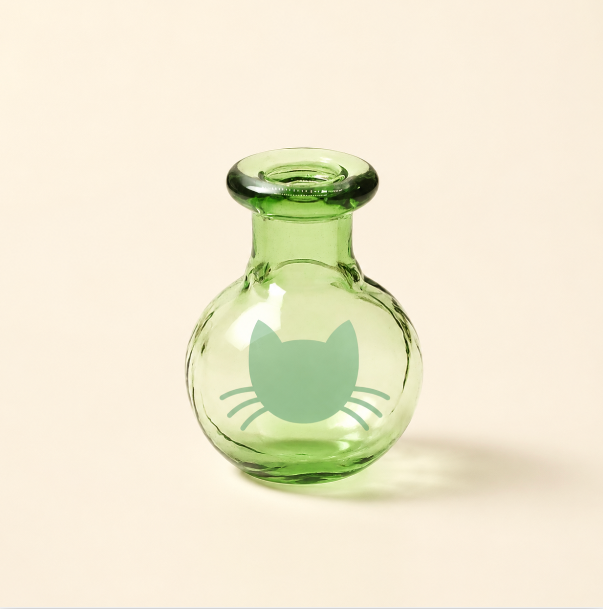 Close-up of a tiny pale green glass Whisker Vase with rounded shape, thick rim, soft cat face graphic, and warm ivory product-page backdrop.