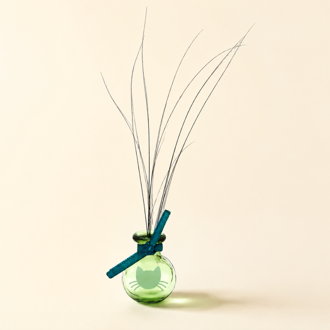 Tiny green glass cat-face Whisker Vase held between fingers with long found whiskers, teal bow, warm ivory background, and refined text overlay.
