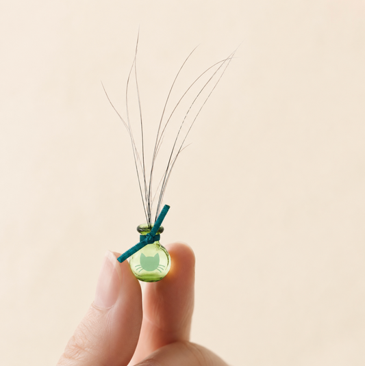 Tiny green glass Whisker Vase with a teal bow and delicate cat whiskers, set against a warm ivory backdrop with soft shadowing and open space.